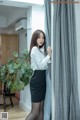 A woman in a white shirt and black skirt leaning against a curtain.