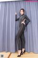 A woman in a black bodysuit posing in front of a curtain.