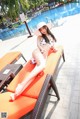 A woman in a white bikini and hat sitting on a lounge chair by a pool.